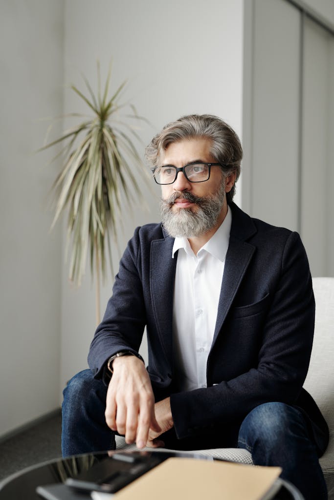 Confident businessman with glasses and beard in a smart suit, sitting in an office.