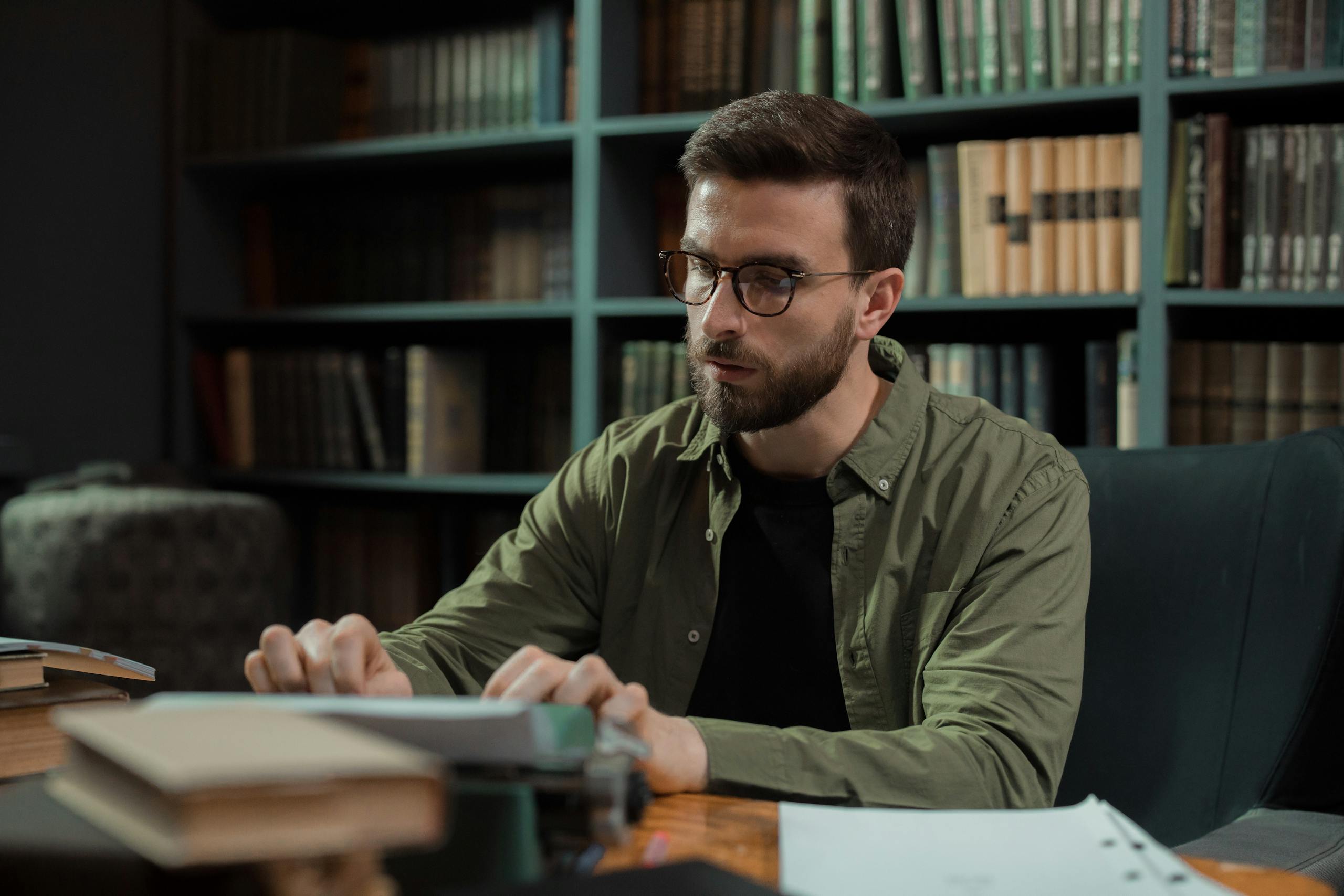 A young man with glasses typing on a vintage typewriter in a library.
