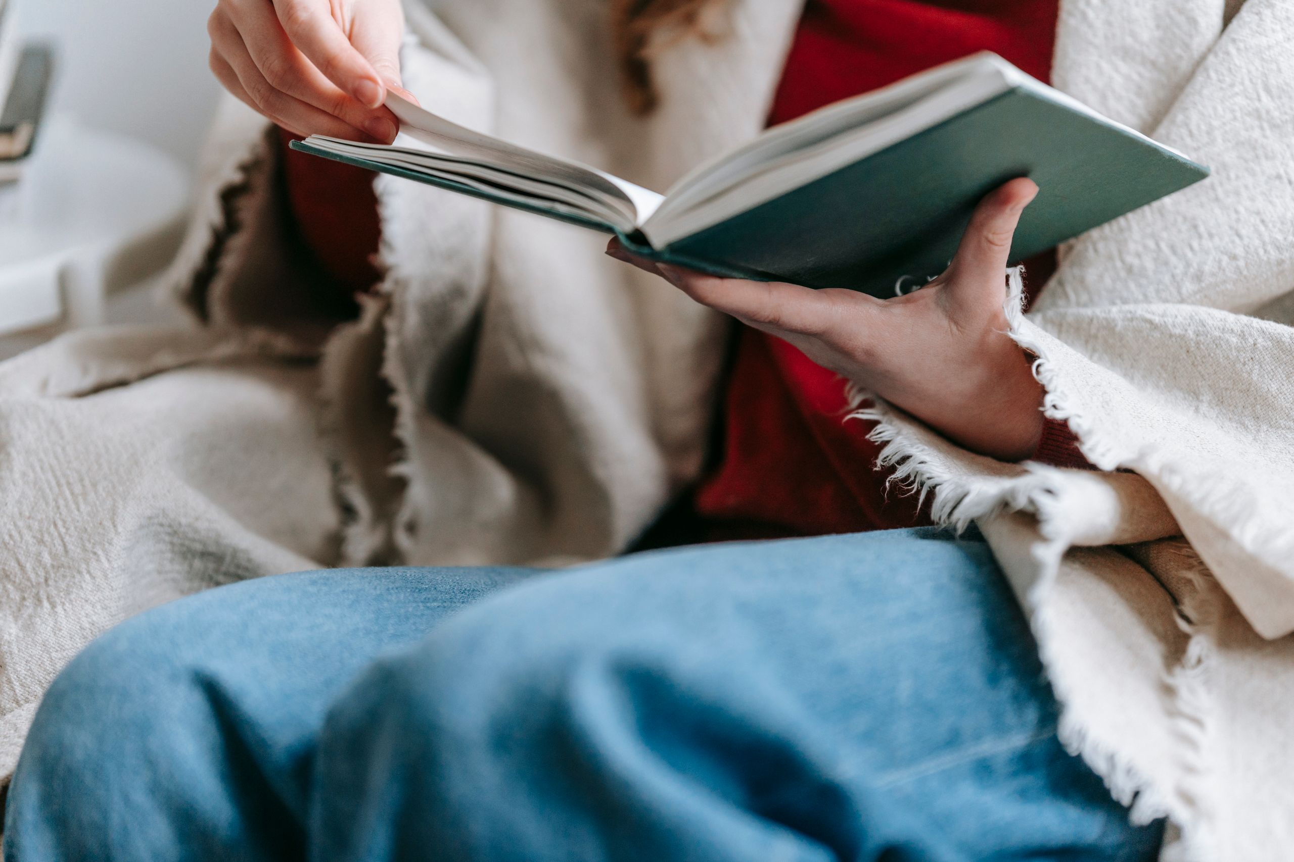 Crop anonymous lady in casual clothes and warm plaid on shoulders reading interesting book while sitting in armchair and spending free time at home